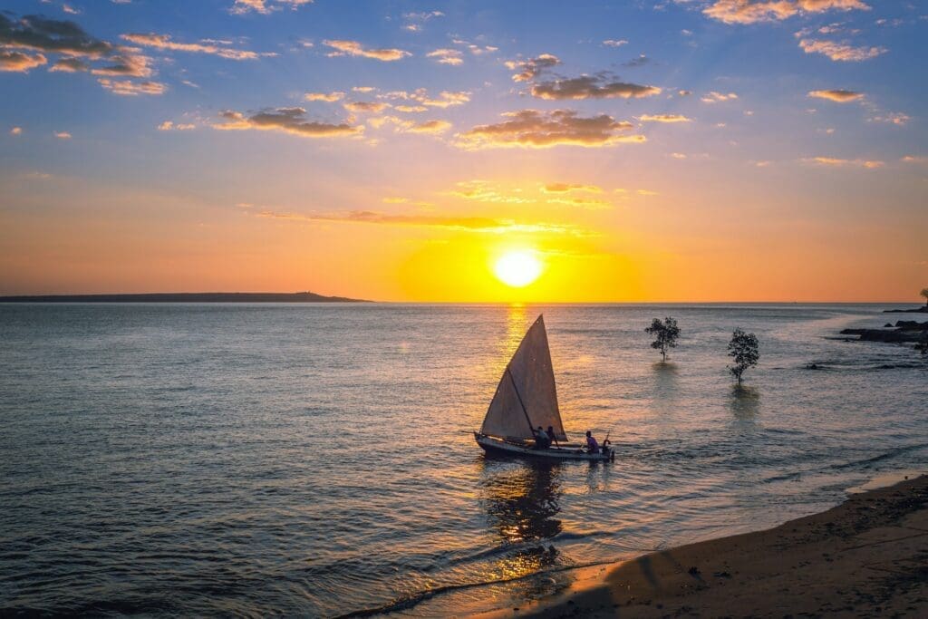Traditional Mozambican dhow sailboat with white canvas sail gliding across calm turquoise water at golden hour, Indian Ocean coastal heritage, editorial travel photography