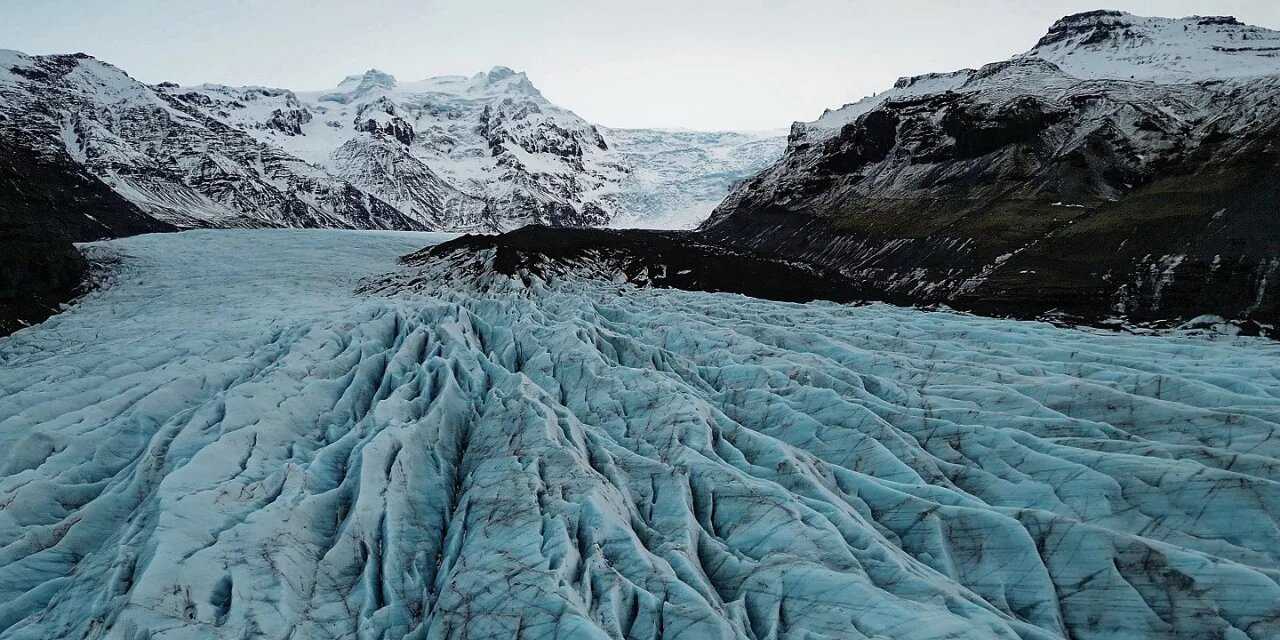 Dramatic Icelandic landscape with a glacial river winding through black volcanic sand toward distant snow-capped mountains under moody grey sky, Iceland honeymoon, editorial travel photography