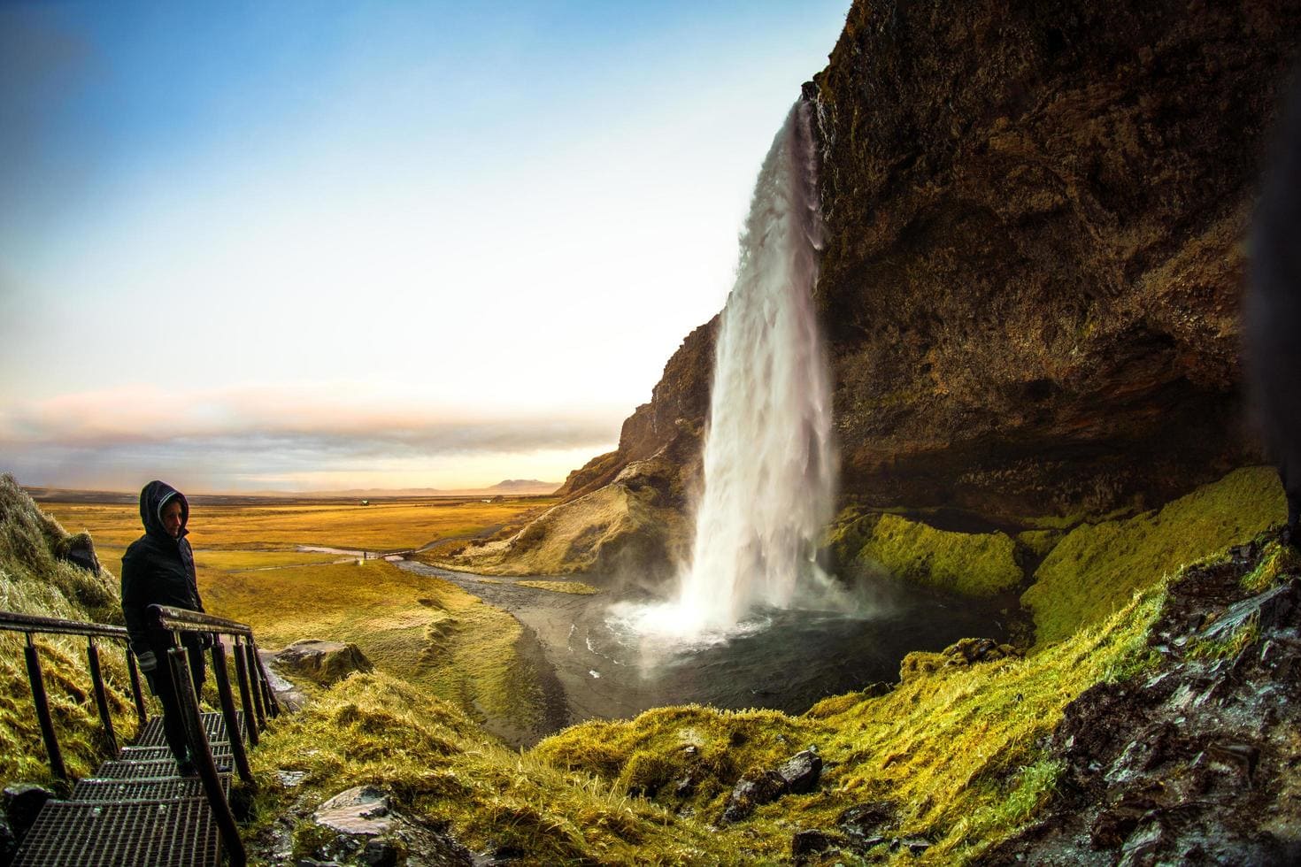 Seljalandsfoss waterfall in Iceland with visitors walking behind the cascade and green moss-covered cliffs visible through the falling water, dramatic honeymoon landscape, editorial photography