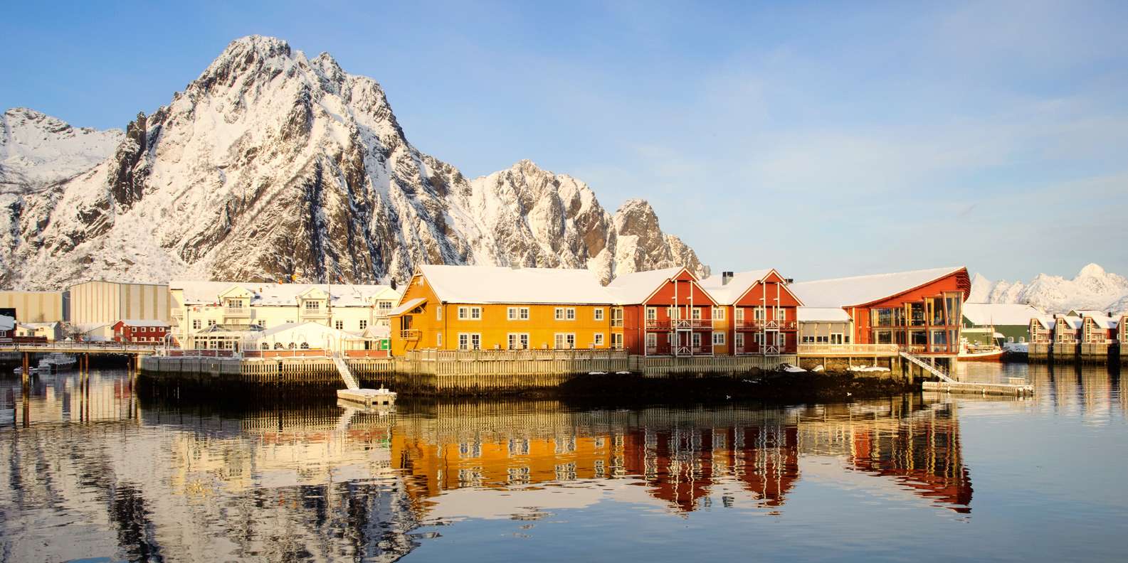 Traditional red rorbuer fishing cabins on stilts over turquoise water in Lofoten Islands Norway with dramatic jagged mountain peaks rising behind, Arctic honeymoon destination, editorial photography