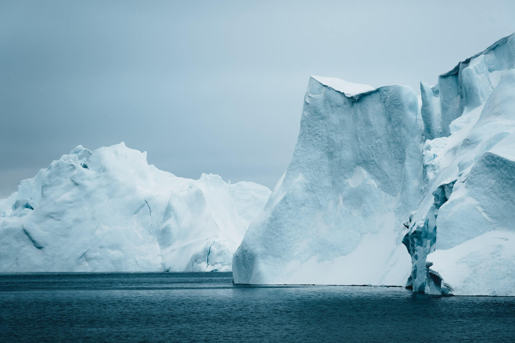 Massive icebergs floating in Ilulissat Icefjord Greenland with deep blue water and distant ice sheet visible under a wide Arctic sky, polar honeymoon destination, editorial travel photography