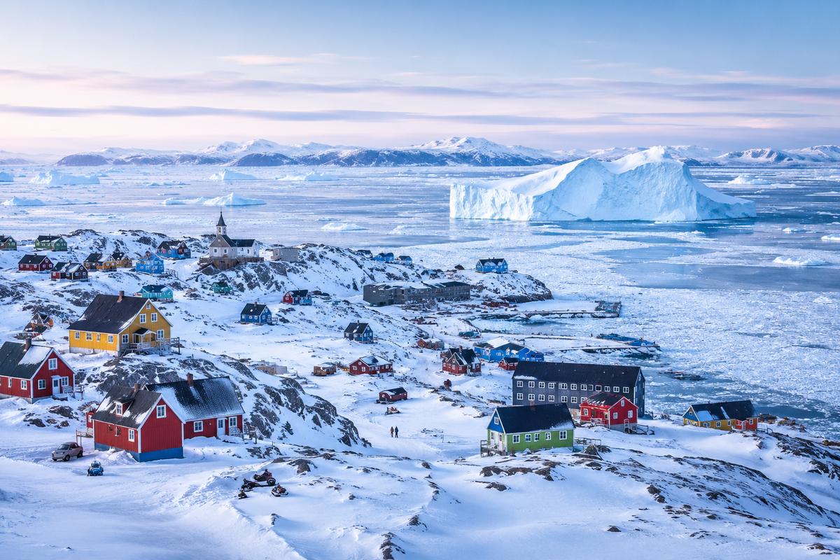 Colourful wooden houses in a small Greenlandic Inuit settlement perched on rocky coastline with icebergs visible in the fjord behind, Arctic cultural honeymoon experience, editorial photography