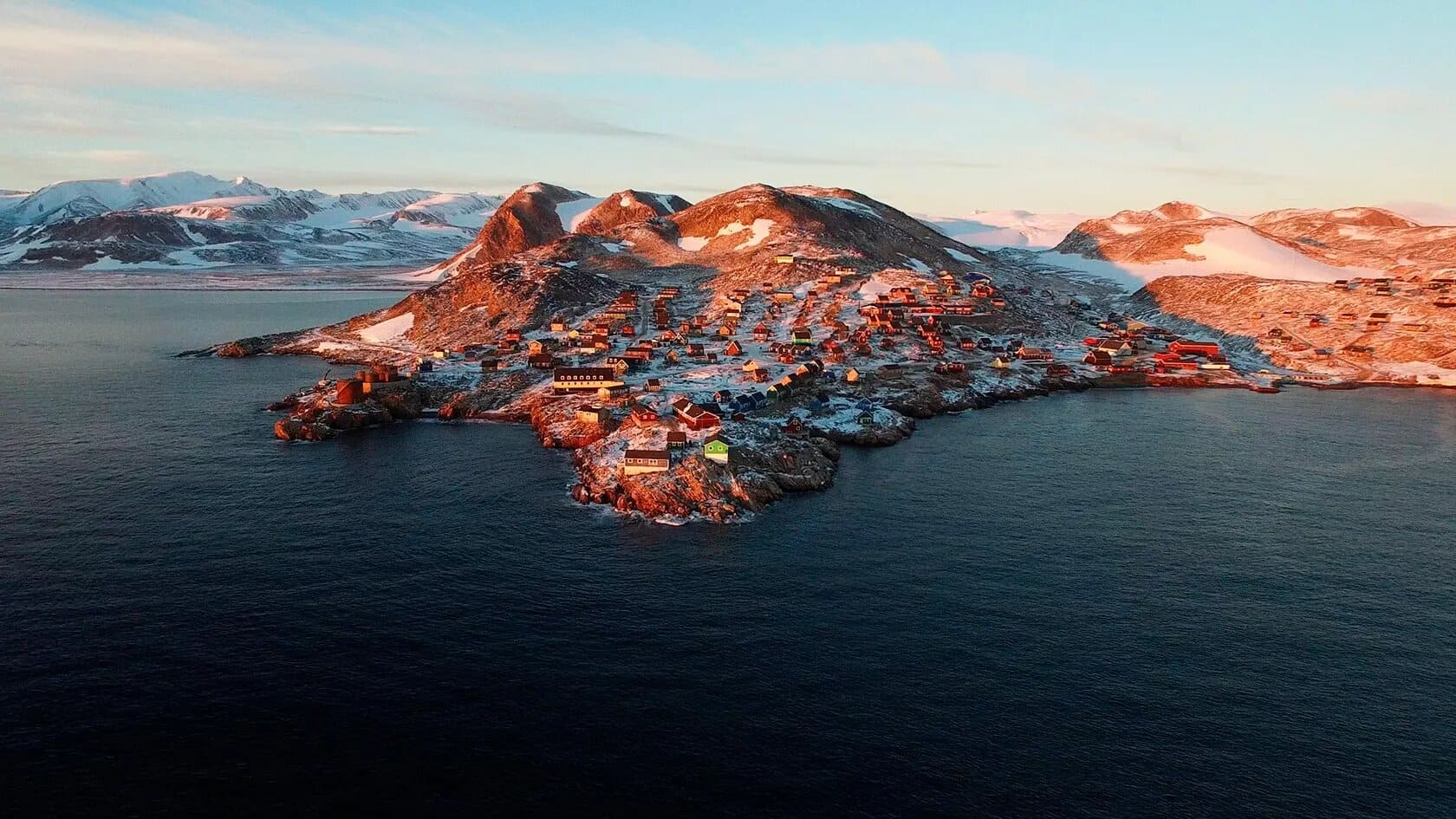 Remote wilderness lodge on a rocky hillside in Greenland overlooking an iceberg-filled fjord at golden hour with vast tundra landscape stretching behind, intimate Arctic honeymoon planning, editorial photography