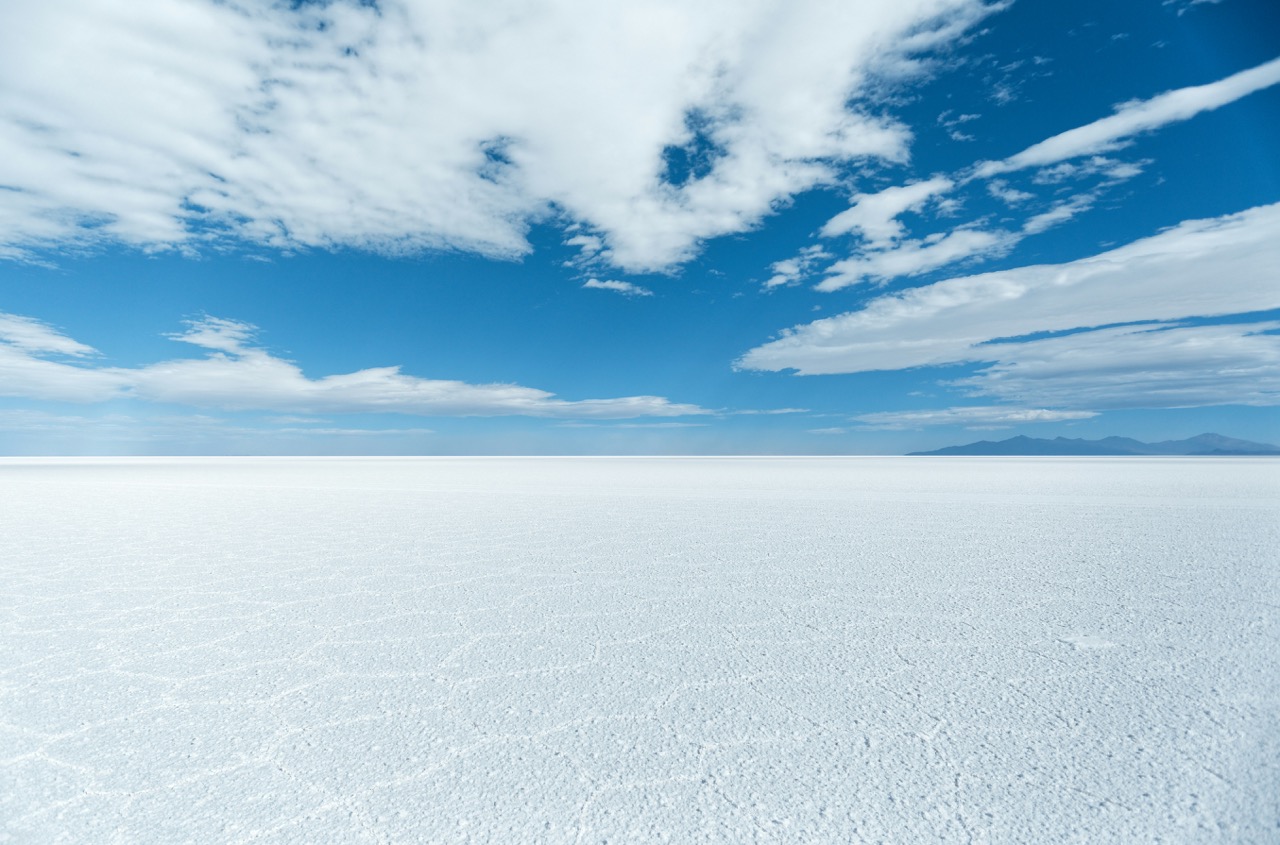 Vast white expanse of Salar de Uyuni salt flat in Bolivia stretching to the horizon under deep blue sky with hexagonal salt patterns in the foreground, surreal honeymoon landscape, editorial travel photography
