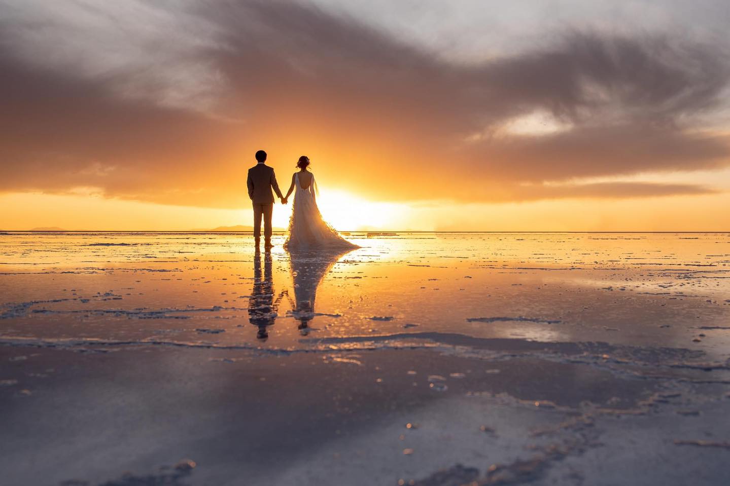 Couple standing on the mirror surface of Salar de Uyuni in Bolivia during wet season with perfect sky reflection creating an infinite horizon effect, surreal romantic honeymoon moment, editorial photography