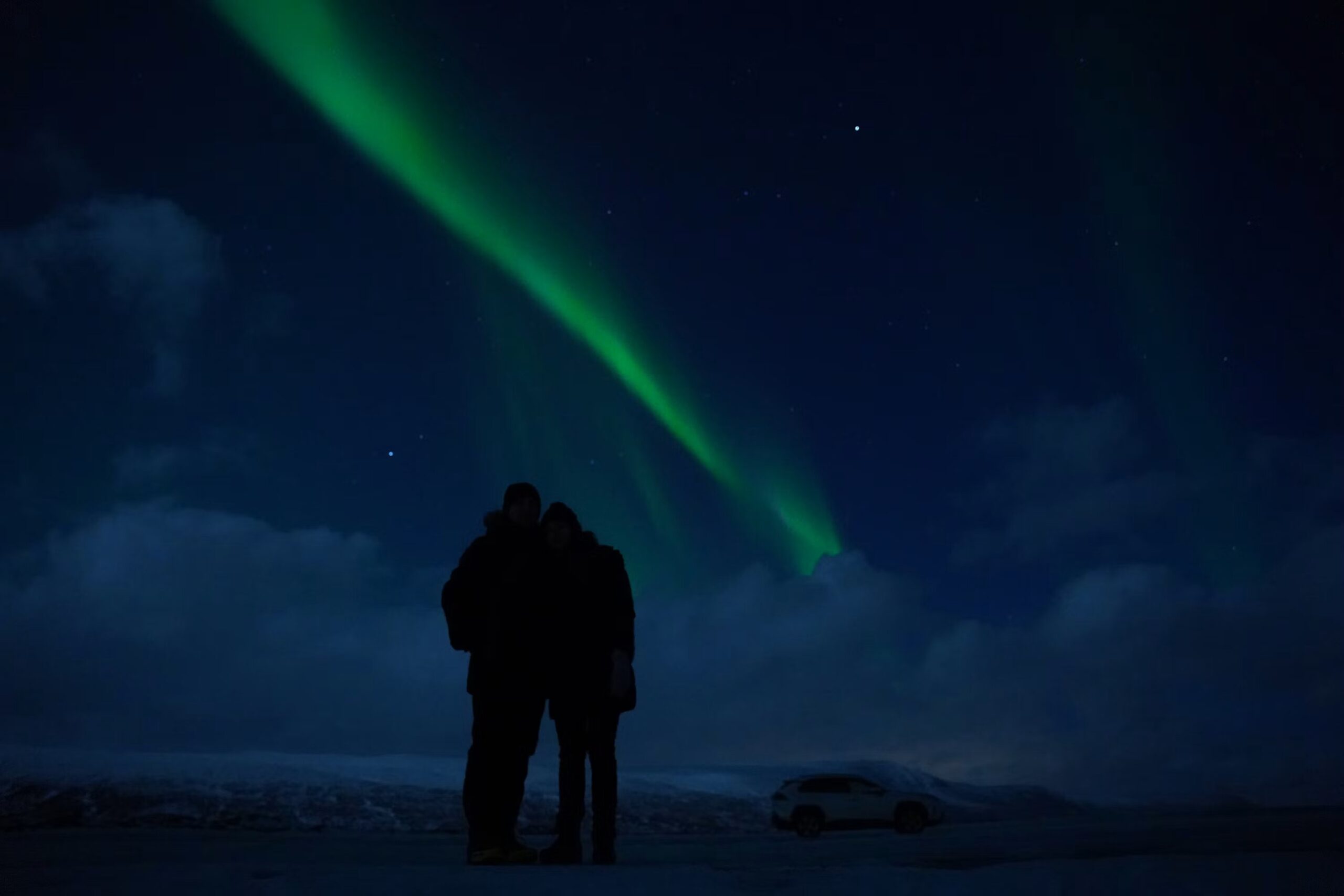 Couple wrapped in blankets on a wooden deck watching a faint green aurora forming on the horizon above a snowy Nordic landscape at night, intimate northern lights honeymoon moment, editorial photography