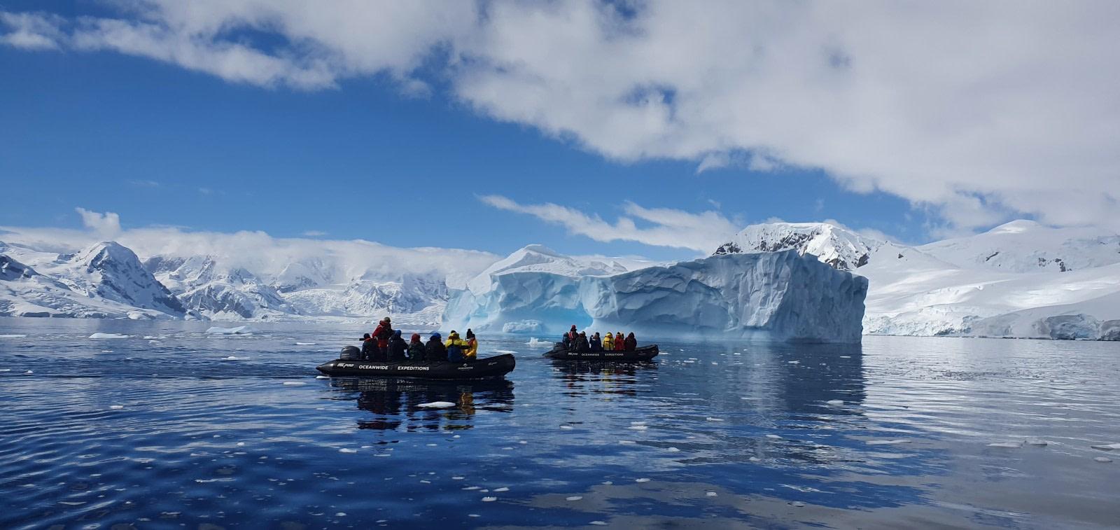 Expedition zodiac approaching a massive iceberg in Antarctic waters with penguins visible on a nearby ice shelf and pale polar light across the scene, adventure honeymoon expedition, editorial photography