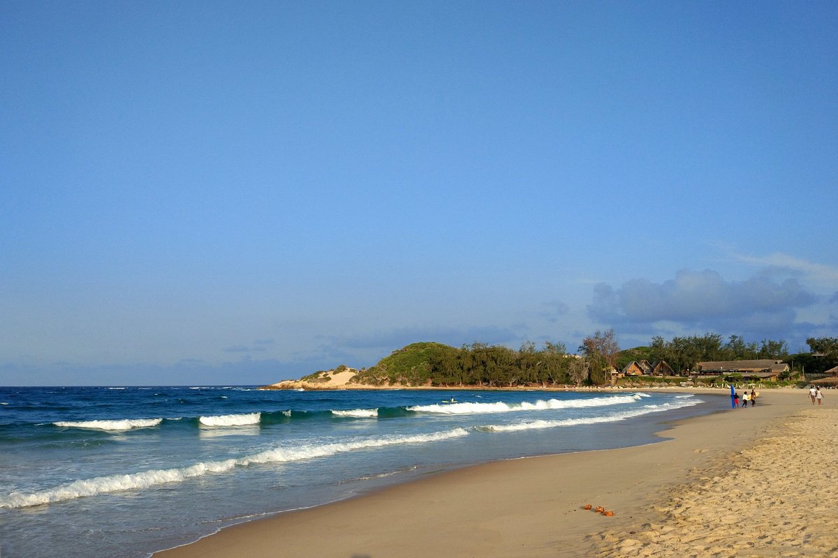Long stretch of empty golden beach at Tofo near Inhambane with gentle turquoise waves and tropical vegetation framing the shoreline, Mozambique surf honeymoon, editorial photography