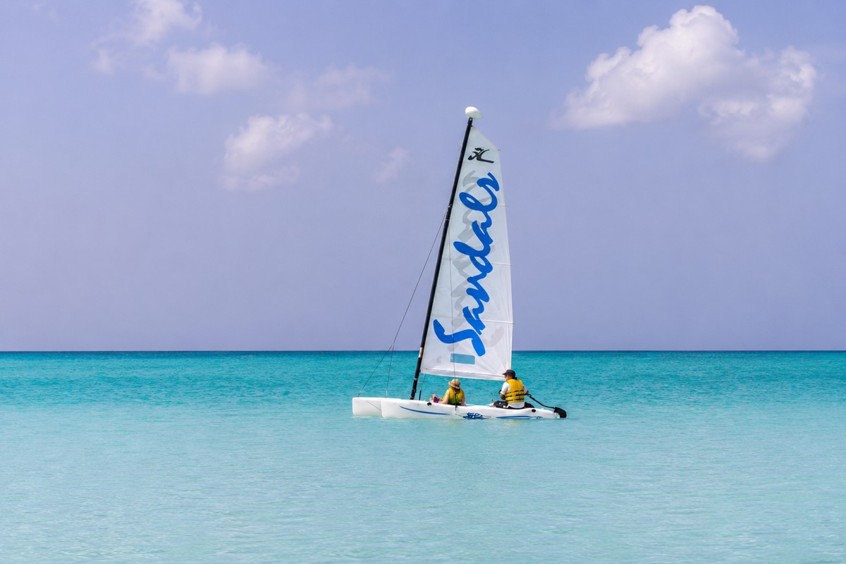 Traditional Caribbean sailing yacht with white sails gliding past Antigua's green coastline at golden hour, warm editorial travel photography for honeymoon content