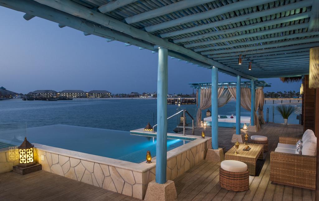 Overwater villa on Banana Island resort Qatar with turquoise Arabian Gulf water and Doha skyline silhouette in the distance at sunset, luxury honeymoon destination, editorial photography
