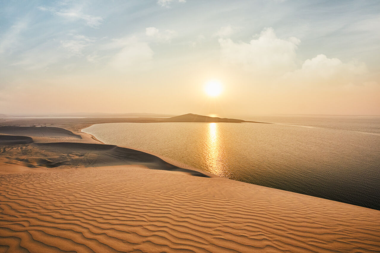 Golden sand dunes of the Qatar desert at sunset with turquoise Inland Sea Khor Al Adaid visible in the distance, Arabian Peninsula landscape, editorial travel photography