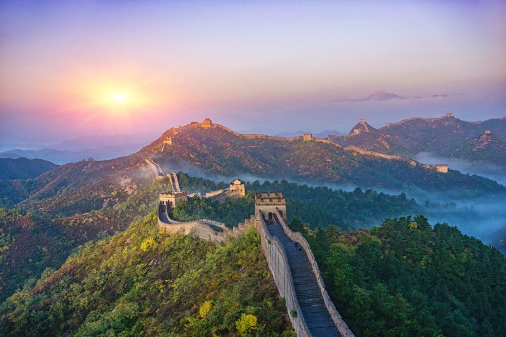 Aerial view of the Great Wall of China winding through forested mountains at golden hour with autumn foliage and distant mist