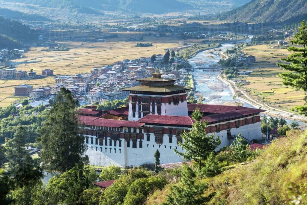 Aerial view of Paro Valley in Bhutan with terraced rice paddies, traditional fortress and prayer flags visible against forest