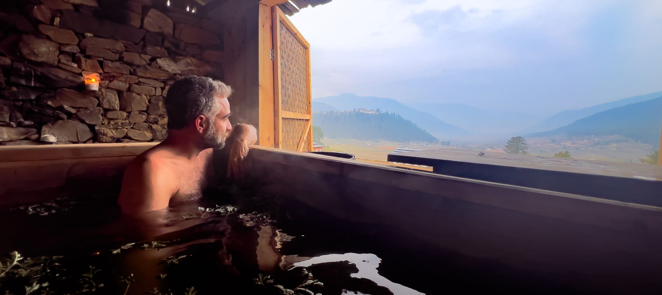 Traditional Bhutanese hot stone bath in a wooden tub with river stones heating in a fire beside a rustic timber bathhouse in a mountain valley, wellness honeymoon experience, editorial photography