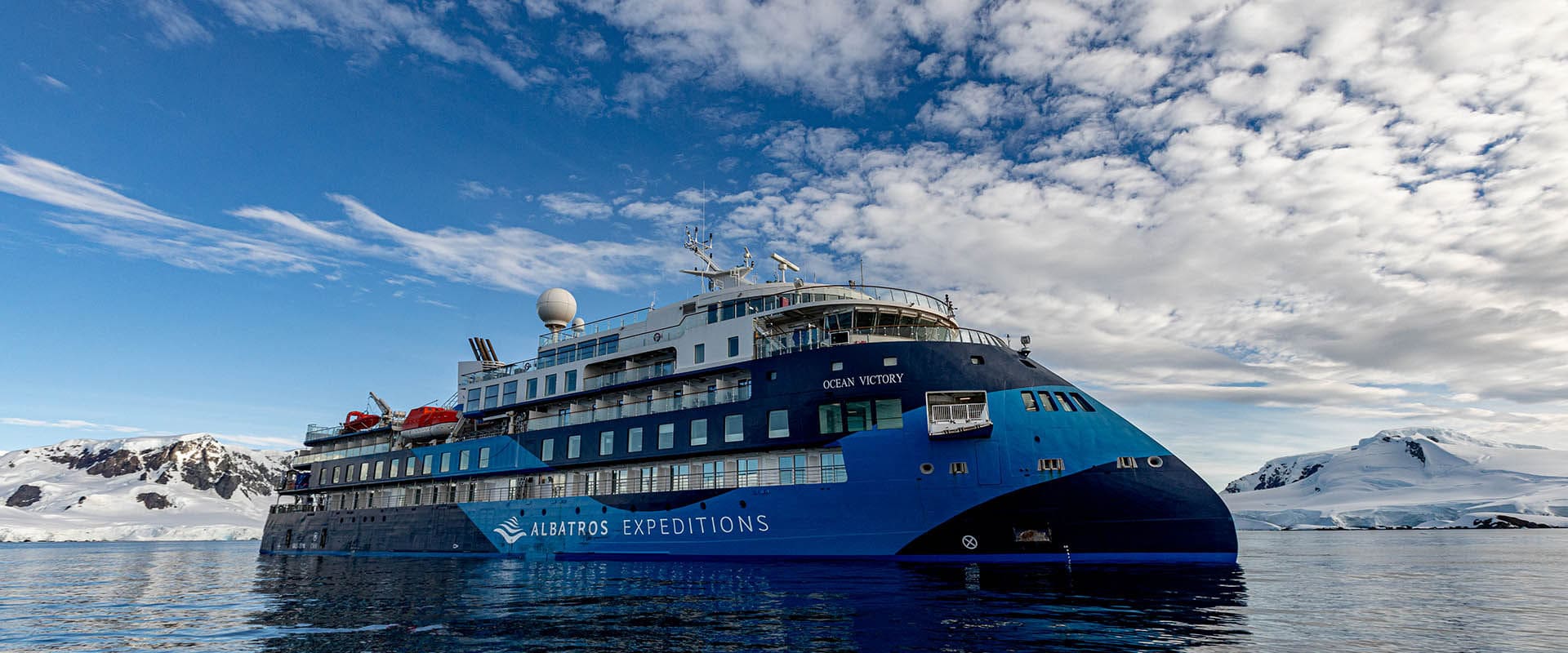 Luxury expedition yacht anchored in a calm Antarctic bay surrounded by snow-covered mountains and floating icebergs under pale blue polar sky, private honeymoon voyage, editorial photography