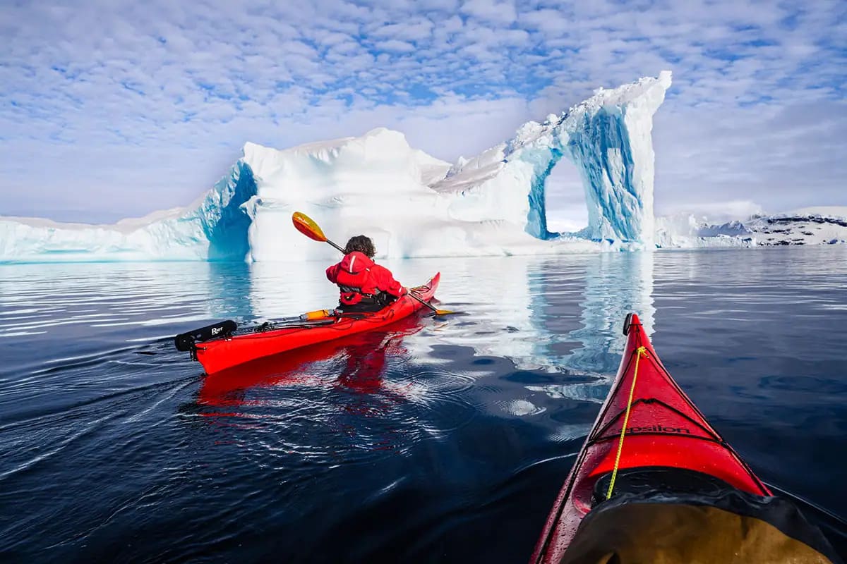 Two sea kayakers paddling between floating icebergs in calm Antarctic waters with glacial cliffs rising behind under soft polar light, adventure honeymoon expedition, editorial photography