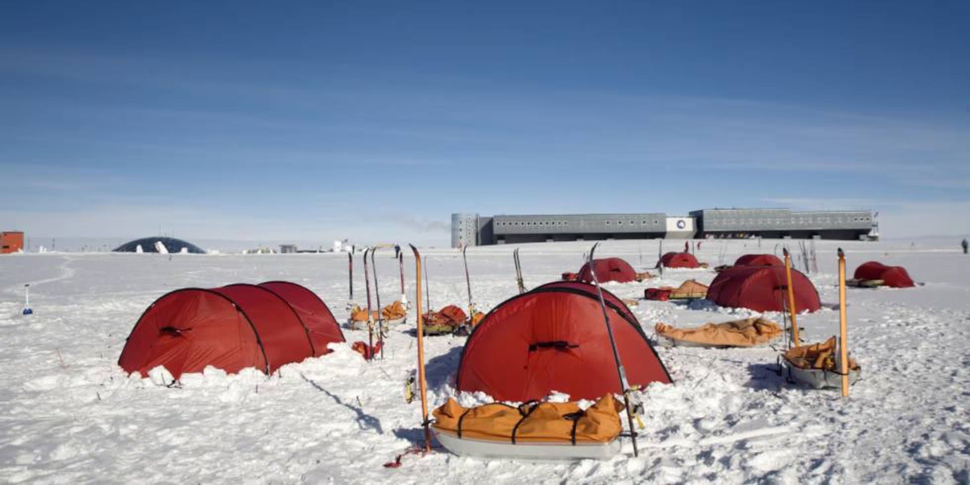 Expedition camp on Antarctic ice shelf at twilight with small tents and distant icebergs under a pale pink and blue polar sky, extreme honeymoon experience, editorial photography