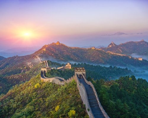 Aerial view of the Great Wall of China winding through forested mountains at golden hour with autumn foliage and distant mist